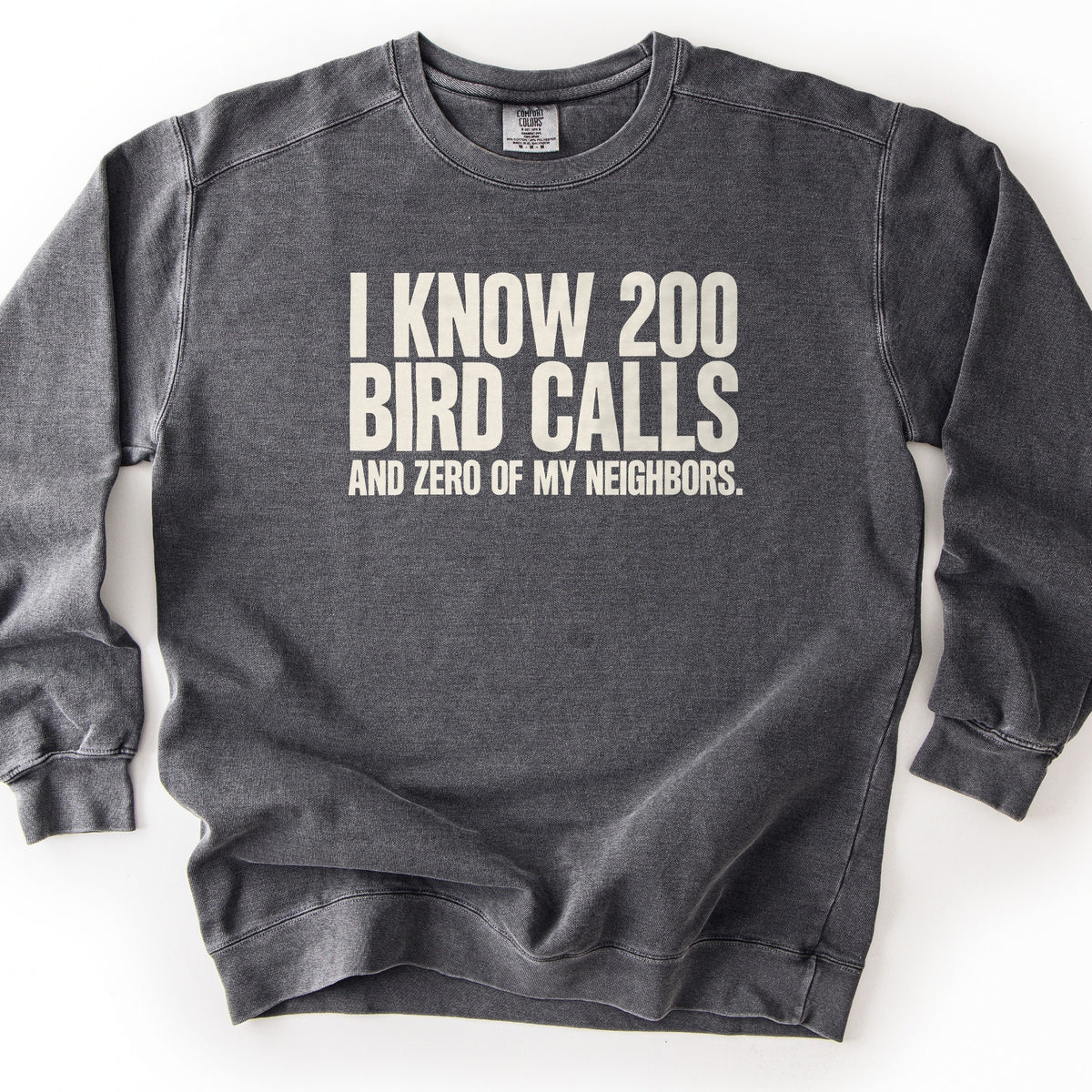 I Know 200 Bird Calls and Zero of My Neighbors Comfort Colors crewneck sweatshirt in Pepper, laid flat. Bold cream capital letters across the chest read I Know 200 Bird Calls, with And Zero of My Neighbors. in smaller letters below. The sweatshirt lies on a plain white background.