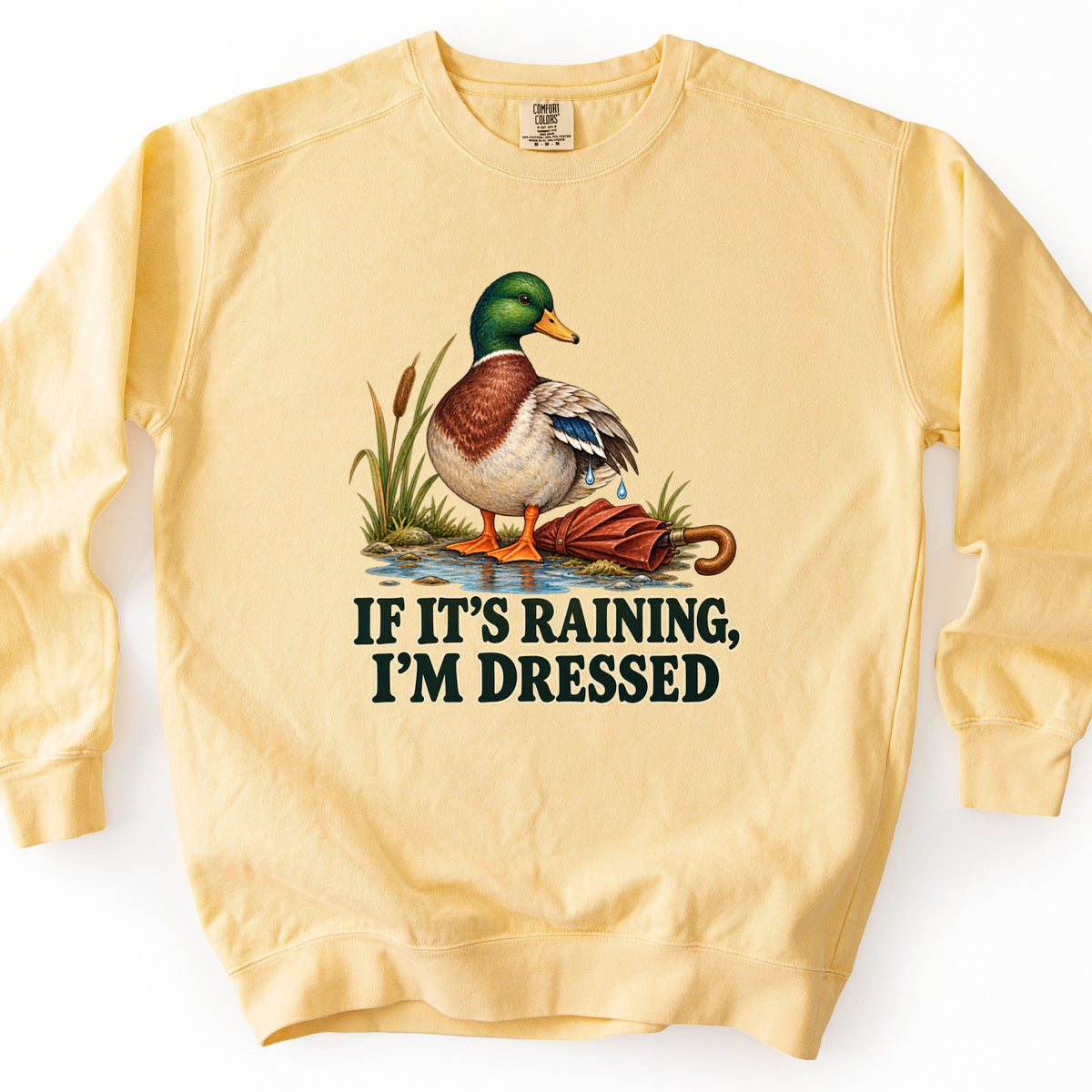 If It's Raining, I'm Dressed Comfort Colors crewneck sweatshirt in Butter, laid flat. A mallard duck with water dripping from its wing stands in a puddle by cattails beside a closed red umbrella, above the words If It's Raining, I'm Dressed in dark green. Shown on a white background.