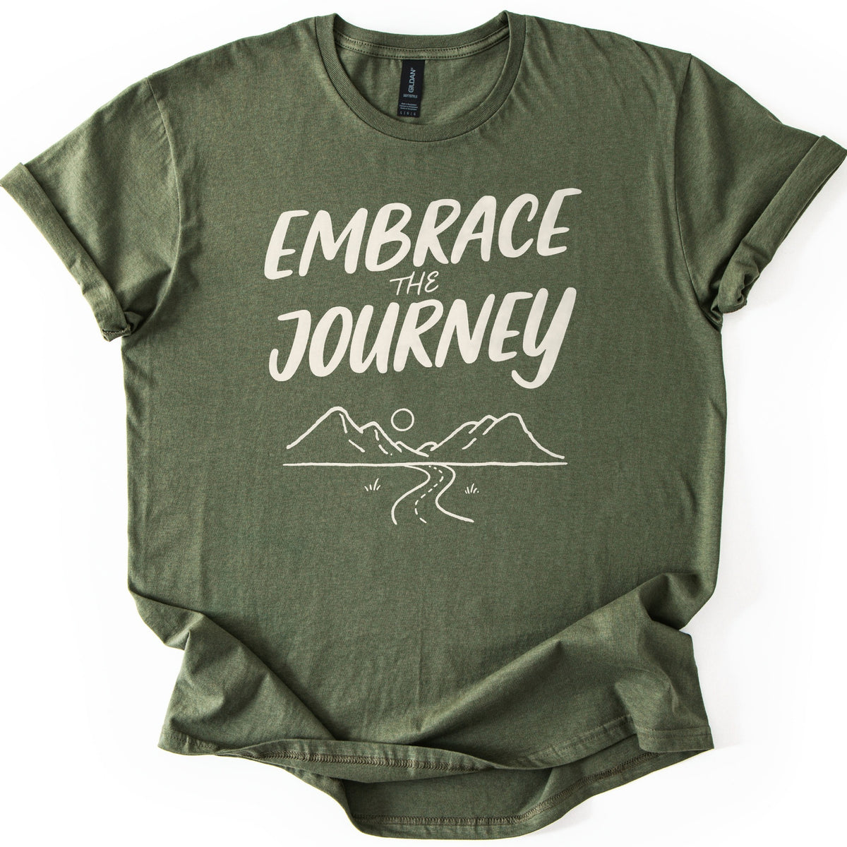 Embrace The Journey Trail Comfort Colors T-shirt in Military Green, laid flat. Cream hand-lettered words Embrace the Journey sit above a line drawing of two mountains, a round sun, and a winding road with tufts of grass. The heathered shirt has cuffed sleeves, on a white background.