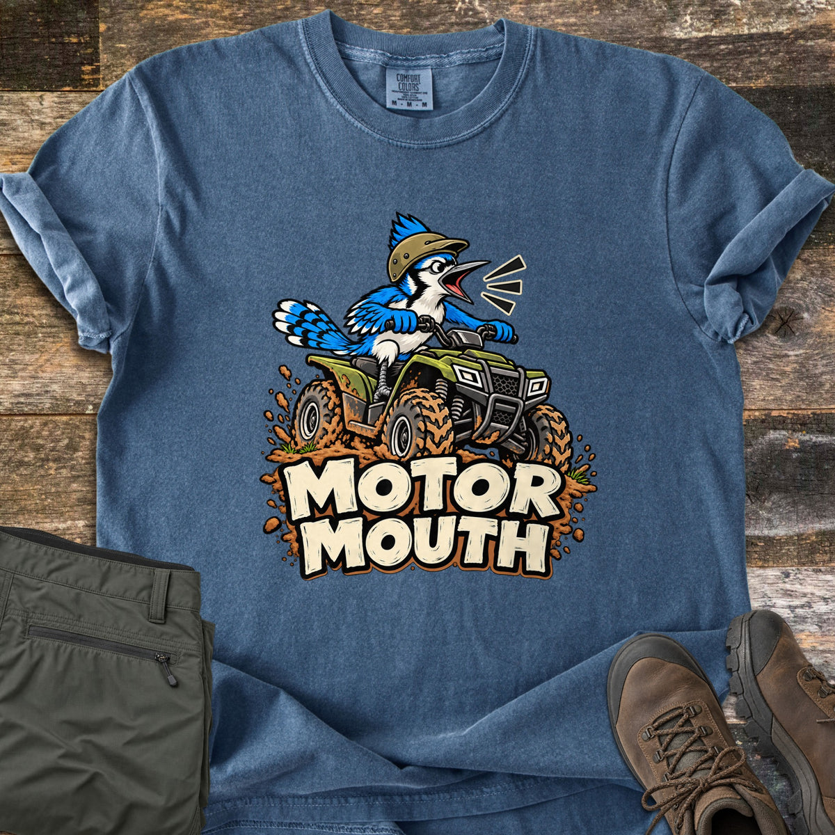 Motor Mouth Comfort Colors T-shirt in Denim, laid flat. A cartoon blue jay in a helmet squawks while riding a green ATV through splashing mud above the words Motor Mouth. Shown on weathered wood with olive pants and brown hiking boots.