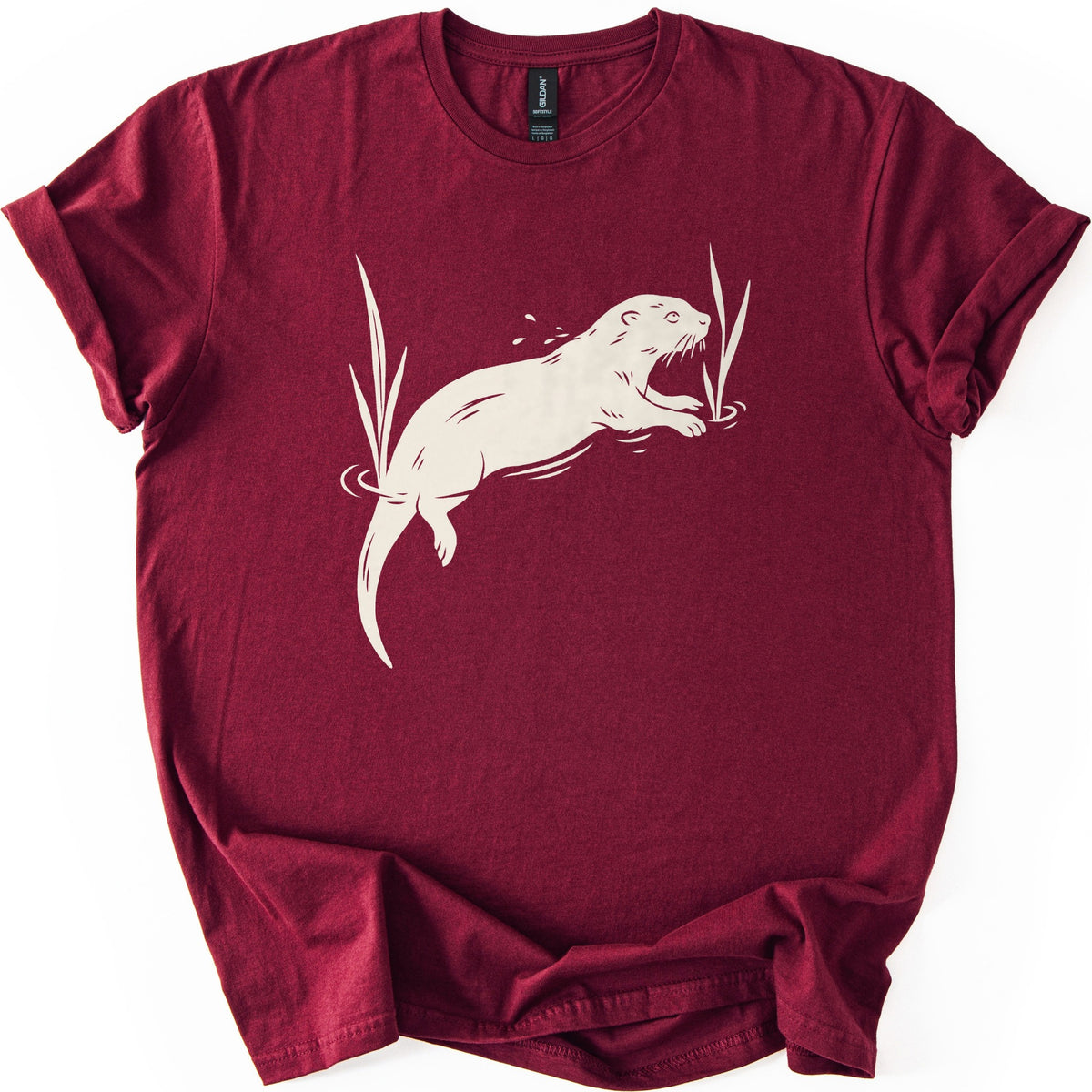 Swimming Otter Comfort Colors T-shirt in Maroon, laid flat. A cream linocut-style otter swims through rippling water between tall reeds, with droplets above its head. No words are printed. The sleeves are cuffed and the shirt lies on a white background.
