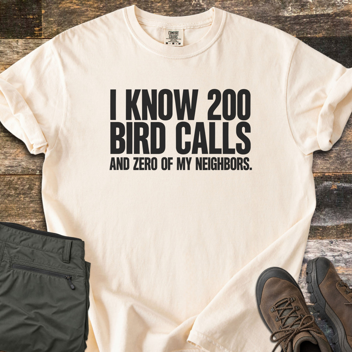 I Know 200 Bird Calls and Zero of My Neighbors Comfort Colors T-shirt in Ivory, laid flat. Bold black capital letters read I KNOW 200 BIRD CALLS AND ZERO OF MY NEIGHBORS. The shirt has rolled sleeves and lies on weathered wood boards beside folded olive hiking pants and brown hiking boots.