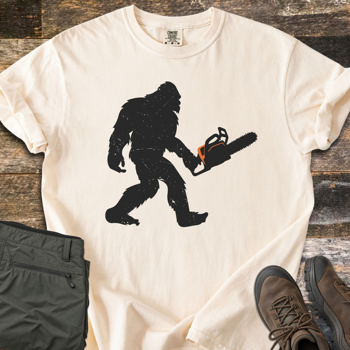 Lumber Squatch Comfort Colors T-shirt in Ivory, laid flat. A distressed black silhouette of Bigfoot walks while carrying a chainsaw with orange accents. The sleeves are rolled, with folded olive pants and brown hiking boots on rustic wood planks.