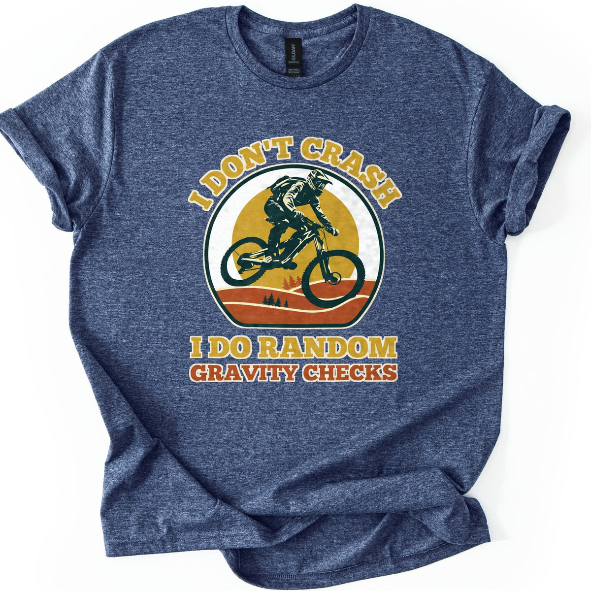 Random Gravity Checks MTB Comfort Colors T-shirt in Heather Navy, laid flat. A mountain biker in a full-face helmet jumps against a mustard sun over orange hills with pine trees, framed by the words I Don't Crash above and I Do Random Gravity Checks below. Sleeves rolled, on white.