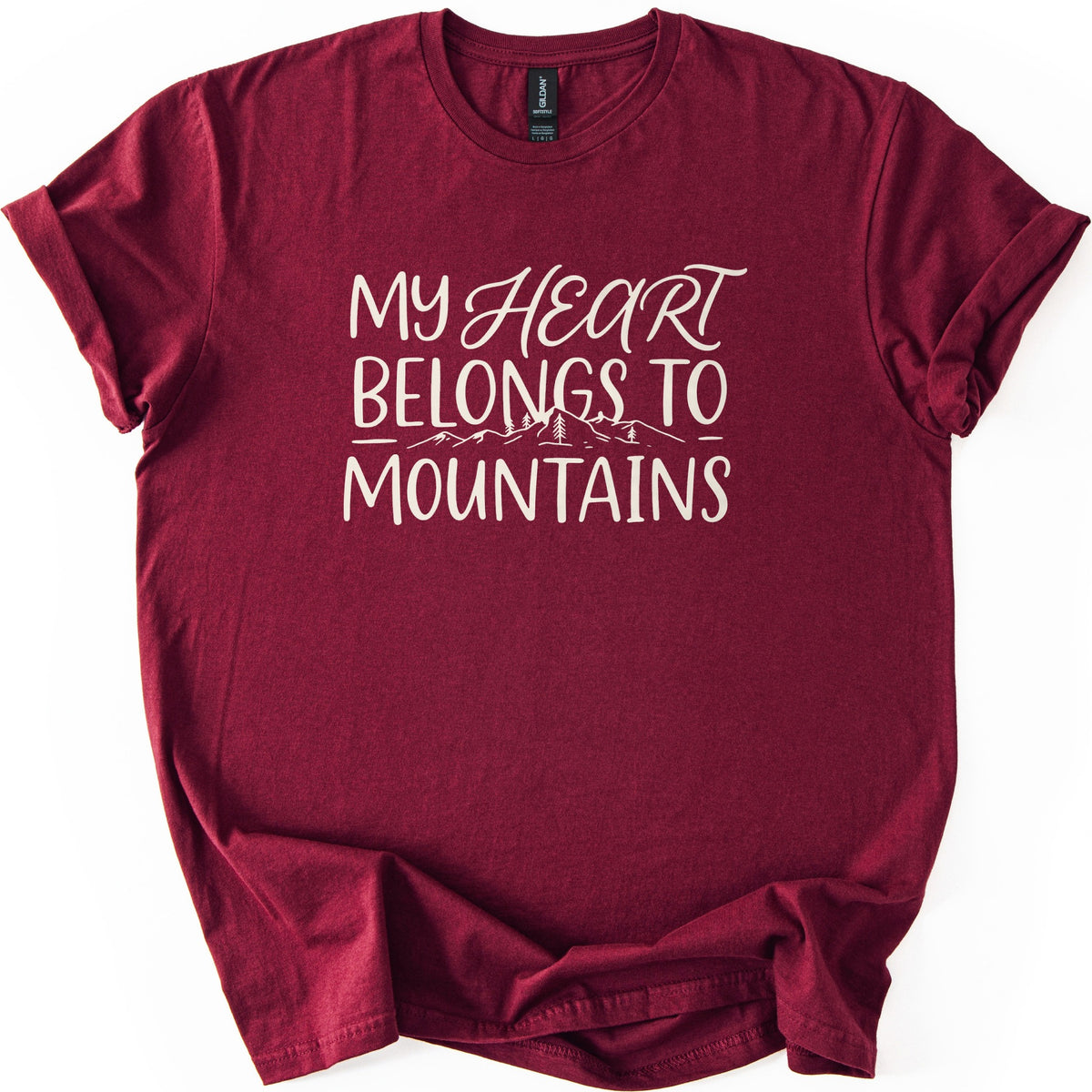 My Heart Belongs to Mountains Comfort Colors T-shirt in Maroon, laid flat. Cream lettering reads My Heart Belongs to Mountains, with Heart in script above a small line drawing of mountains and pine trees. The shirt has cuffed sleeves and lies on a white background.