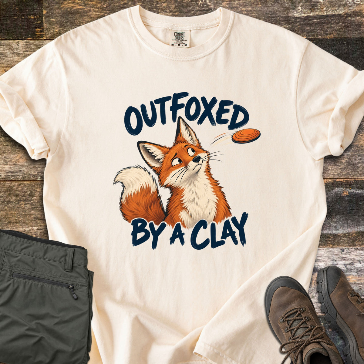 Outfoxed by a Clay Comfort Colors T-shirt in Ivory, laid flat. A cartoon red fox glances sideways at an orange clay target flying past, between the words Outfoxed By A Clay in navy brush letters. Sleeves rolled on rustic wood with olive pants and brown hiking boots.