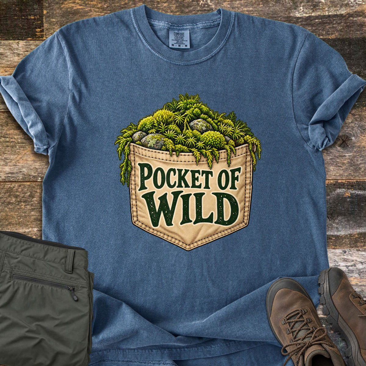 Pocket of Wild Comfort Colors T-shirt in Denim, laid flat. A tan denim back pocket overflowing with green moss, ferns and lichen-covered stones, with the words Pocket of Wild printed on it. The shirt lies on weathered wood beside olive hiking pants and brown hiking boots.
