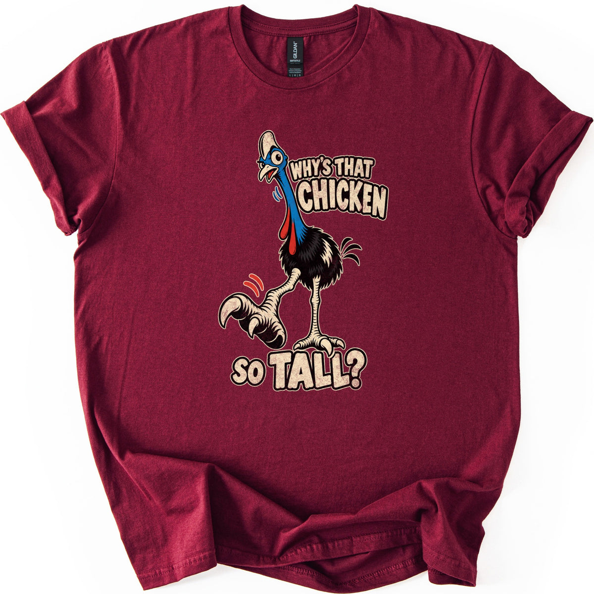 Why's That Chicken So Tall Comfort Colors T-shirt in Maroon, laid flat. A cartoon cassowary with a blue head, red wattles and long pale legs raises a big clawed foot next to the words Why's That Chicken So Tall? The shirt lies on a white surface with rolled sleeves.