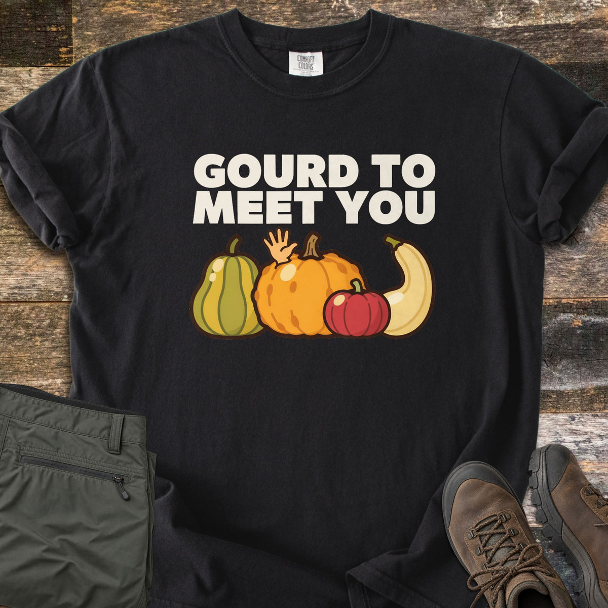 Gourd To Meet You Comfort Colors T-shirt in Black, laid flat. Off-white words Gourd To Meet You above cartoon gourds: a green striped squash, an orange pumpkin with a waving hand, a red pumpkin and a cream squash. Olive pants and brown hiking boots sit on wood planks.