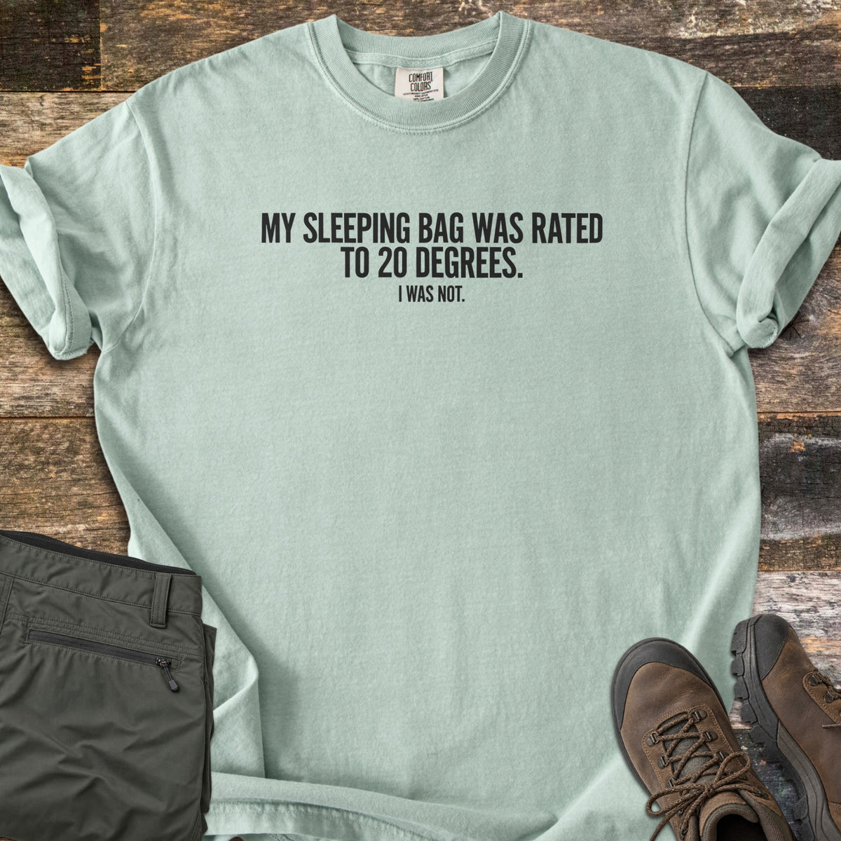 Rated To 20 Degrees Comfort Colors T-shirt in Bay, laid flat. Black text across the chest reads My Sleeping Bag Was Rated To 20 Degrees. I Was Not. The shirt has rolled sleeves and lies on weathered wood planks beside folded olive hiking pants and brown hiking boots.