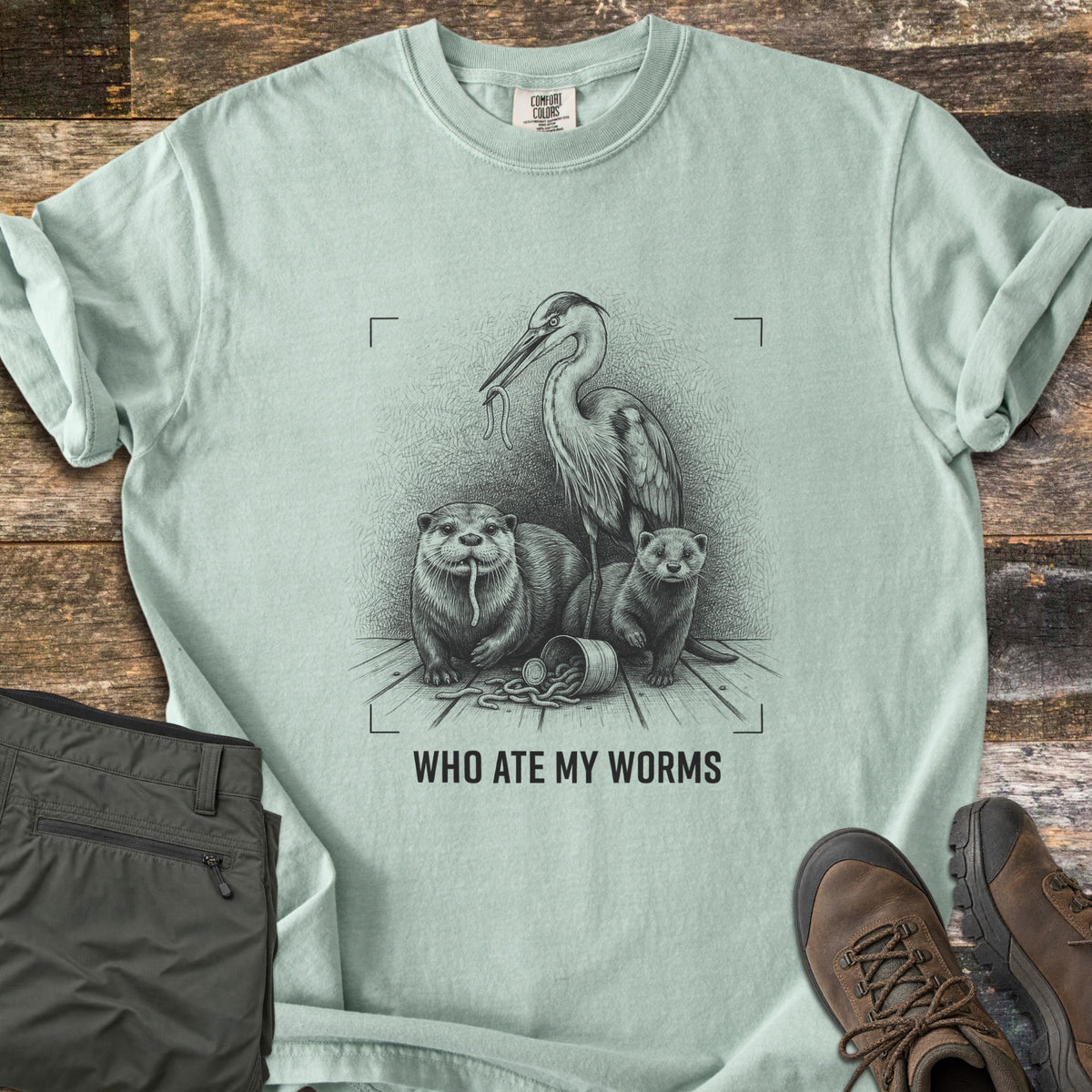 Who Ate My Worms Comfort Colors T-shirt in Bay, laid flat. A black sketch of a heron and an otter each eating a worm beside a mink and a spilled bait can, inside viewfinder corners, above the words Who Ate My Worms. Olive pants and brown hiking boots lie on weathered wood.
