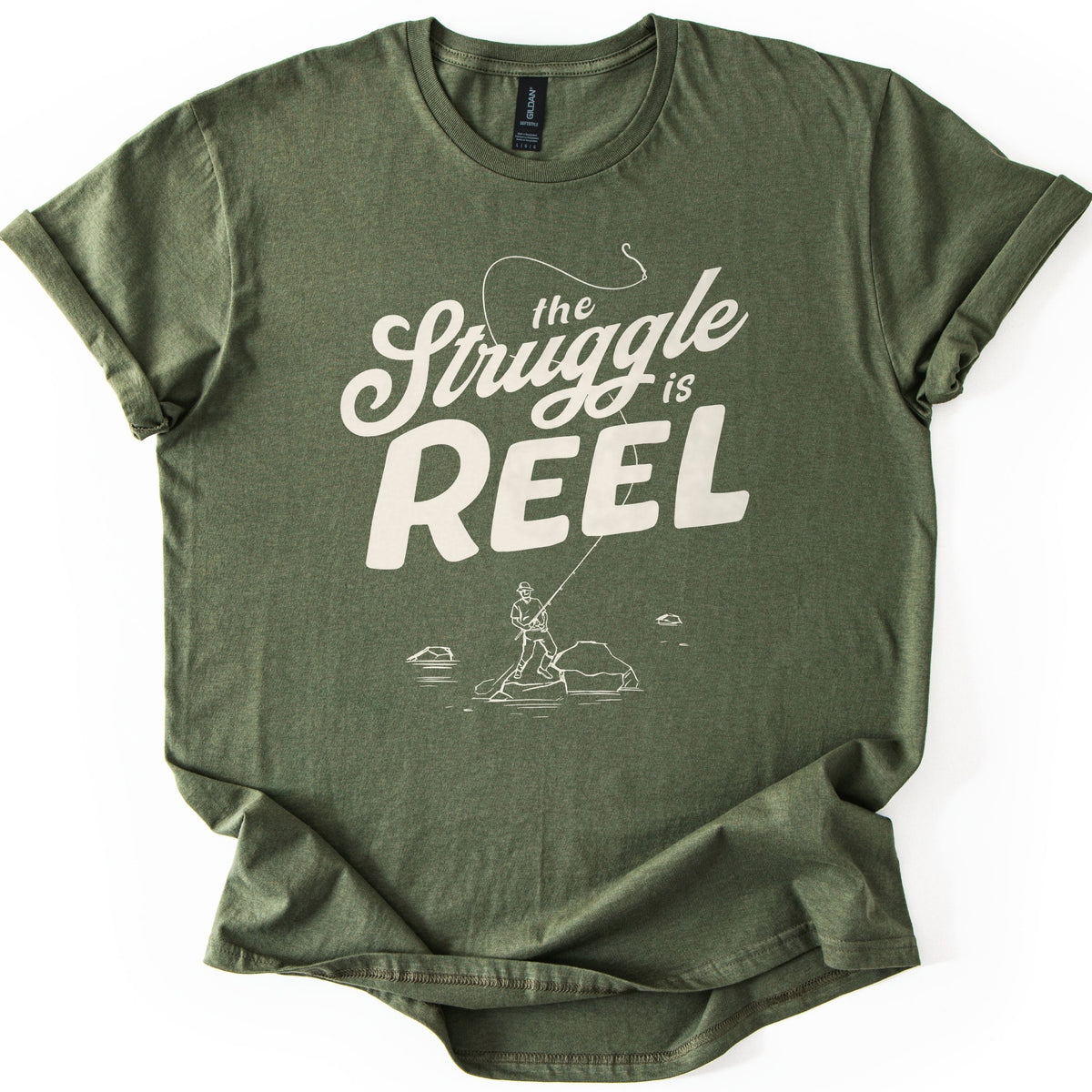 Struggle Is Reel Angler Comfort Colors T-shirt in Military Green, laid flat. Cream text reads the Struggle is REEL, with a fishing line arcing over the words to a hook. Below, a line drawing of an angler in a hat casts from a rock in the water. Sleeves cuffed, on a white background.