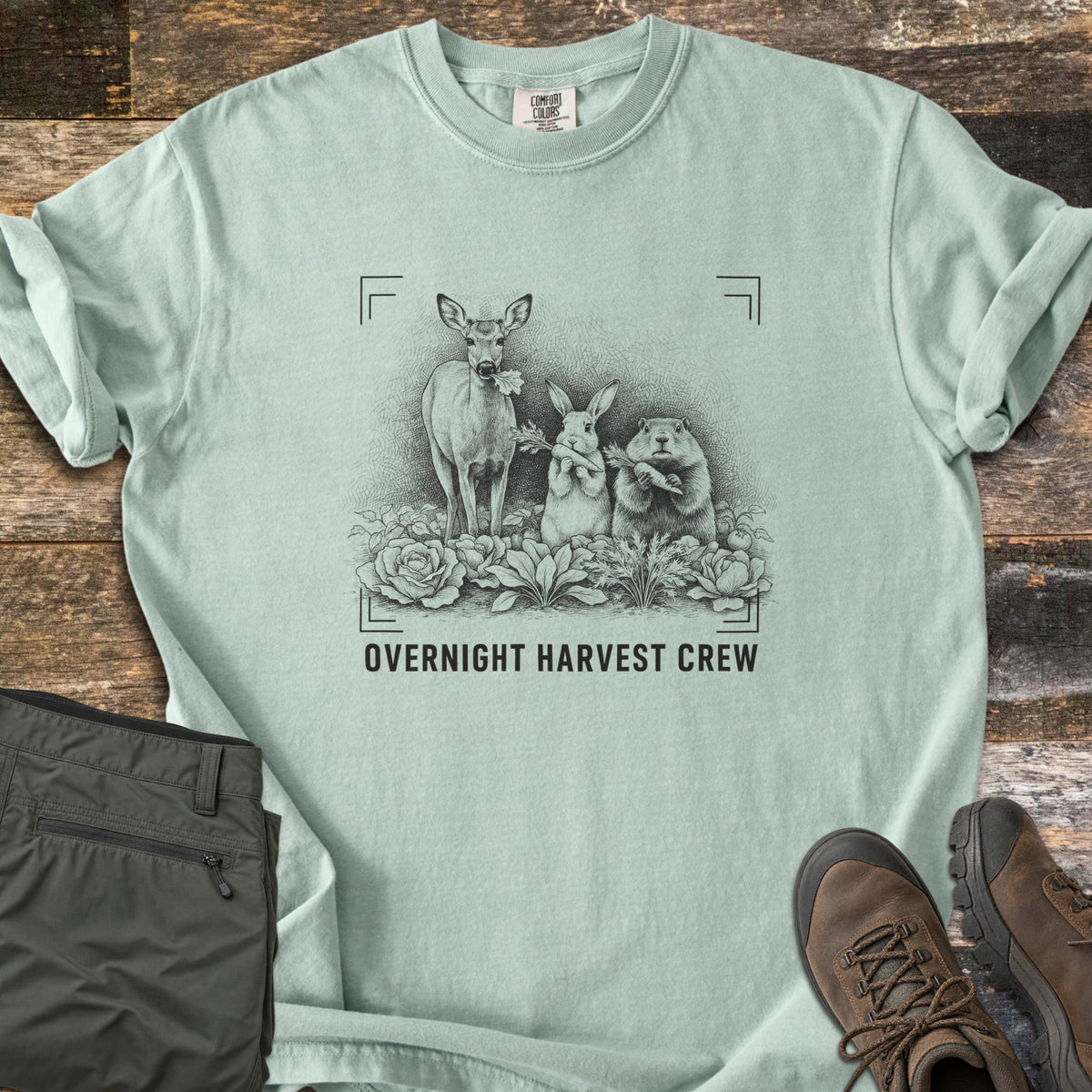Overnight Harvest Crew Comfort Colors T-shirt in Bay, laid flat. A dark sketch of a deer chewing a leaf, a rabbit and a groundhog holding carrots in a cabbage patch, inside camera frame corners above the words Overnight Harvest Crew. Olive pants and brown boots lie on wood.