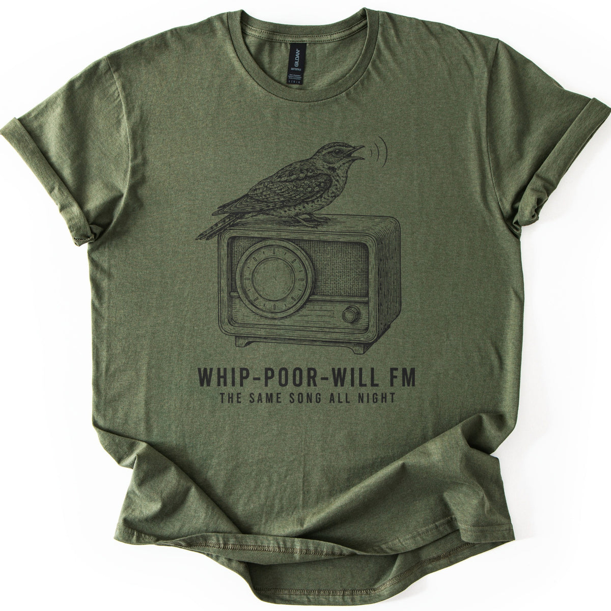 Whip-Poor-Will FM Comfort Colors T-shirt in Military Green, laid flat. A black engraving-style bird sings atop a vintage tabletop radio above the words WHIP-POOR-WILL FM and THE SAME SONG ALL NIGHT. The shirt lies on a white surface with rolled sleeves.