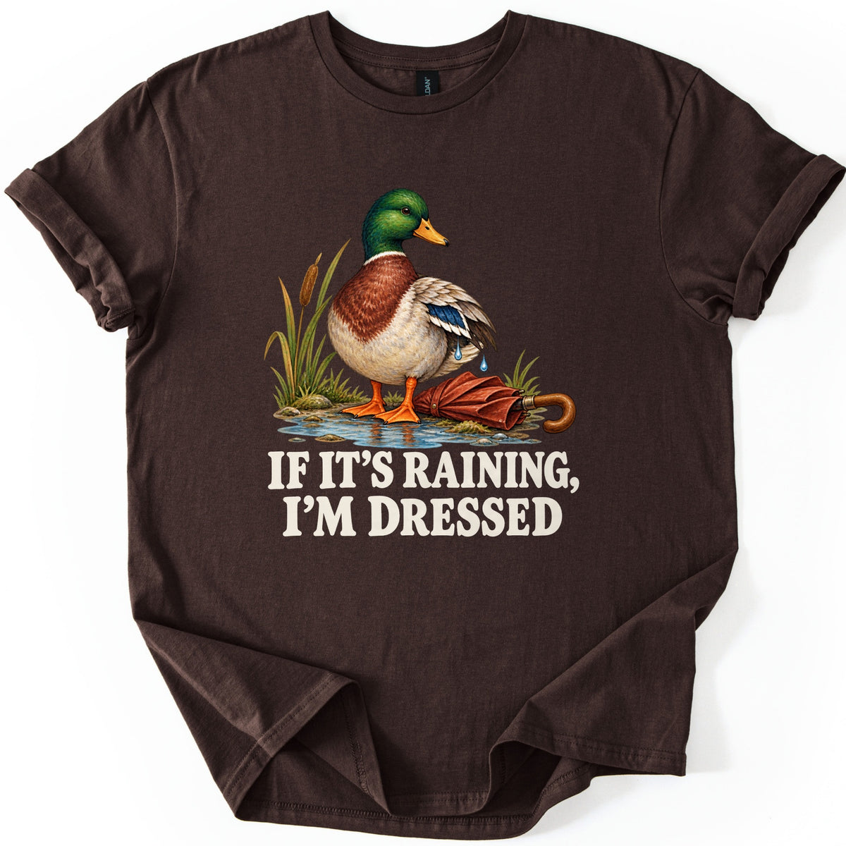 If It's Raining, I'm Dressed Comfort Colors T-shirt in Dark Chocolate, laid flat. A mallard duck with water dripping off its wing stands in a puddle by cattails beside a closed red umbrella, above the words IF IT'S RAINING, I'M DRESSED in cream. Cuffed sleeves, white background.