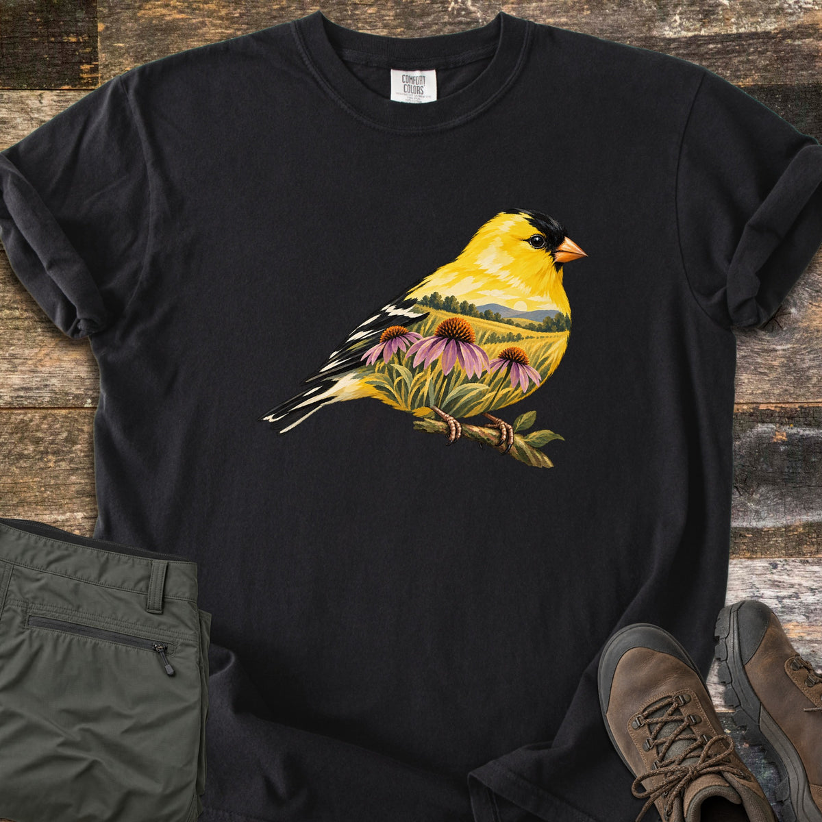 Goldfinch Meadow Comfort Colors T-shirt in Black, laid flat. A bright yellow goldfinch on a twig, its body holding a meadow of purple coneflowers, hills and a low sun. No text. Sleeves rolled, next to olive cargo pants and brown hiking boots on weathered wood boards.