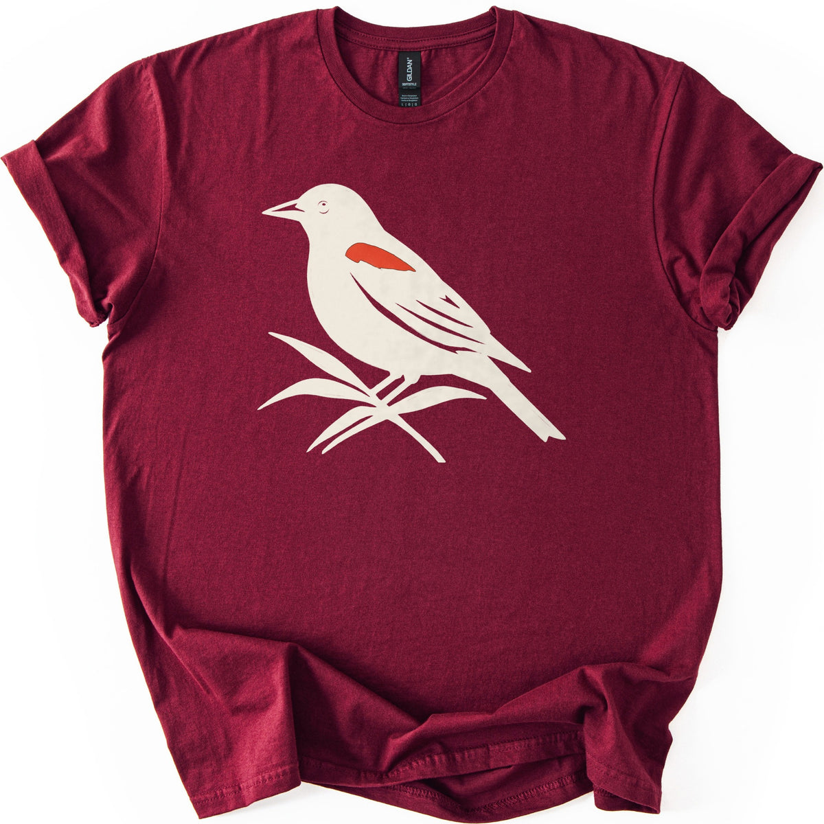 Red Winged Blackbird Comfort Colors T-shirt in Maroon, laid flat. A cream silhouette of a red-winged blackbird with a red shoulder patch, perched on slender leaves, with no words. The shirt has rolled sleeves and lies on a white background.