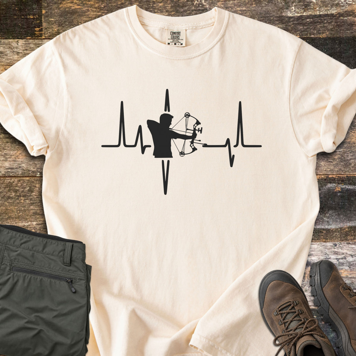 Compound Bow Heartbeat Comfort Colors T-shirt in Ivory, laid flat. A black heartbeat line with the silhouette of an archer drawing a compound bow at its center. Sleeves are rolled on a wood plank floor, with olive cargo pants and brown hiking boots beside it.