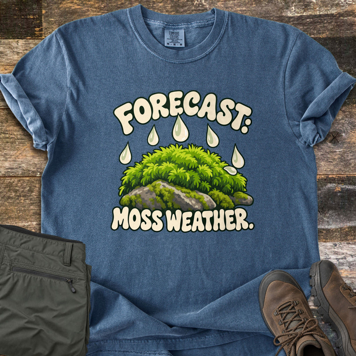 Forecast Moss Weather Comfort Colors T-shirt in Denim, laid flat. A green mound of moss on gray rocks under falling raindrops, with Forecast: above and Moss Weather. below in cream lettering. Olive hiking pants and brown hiking boots sit beside it on weathered wood planks.