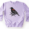 Red Winged Blackbird Comfort Colors crewneck sweatshirt in Orchid, laid flat. A black silhouette of a red-winged blackbird with a red shoulder patch perches on slender leafy stems across the chest. No words are printed. The sweatshirt lies on a white surface.