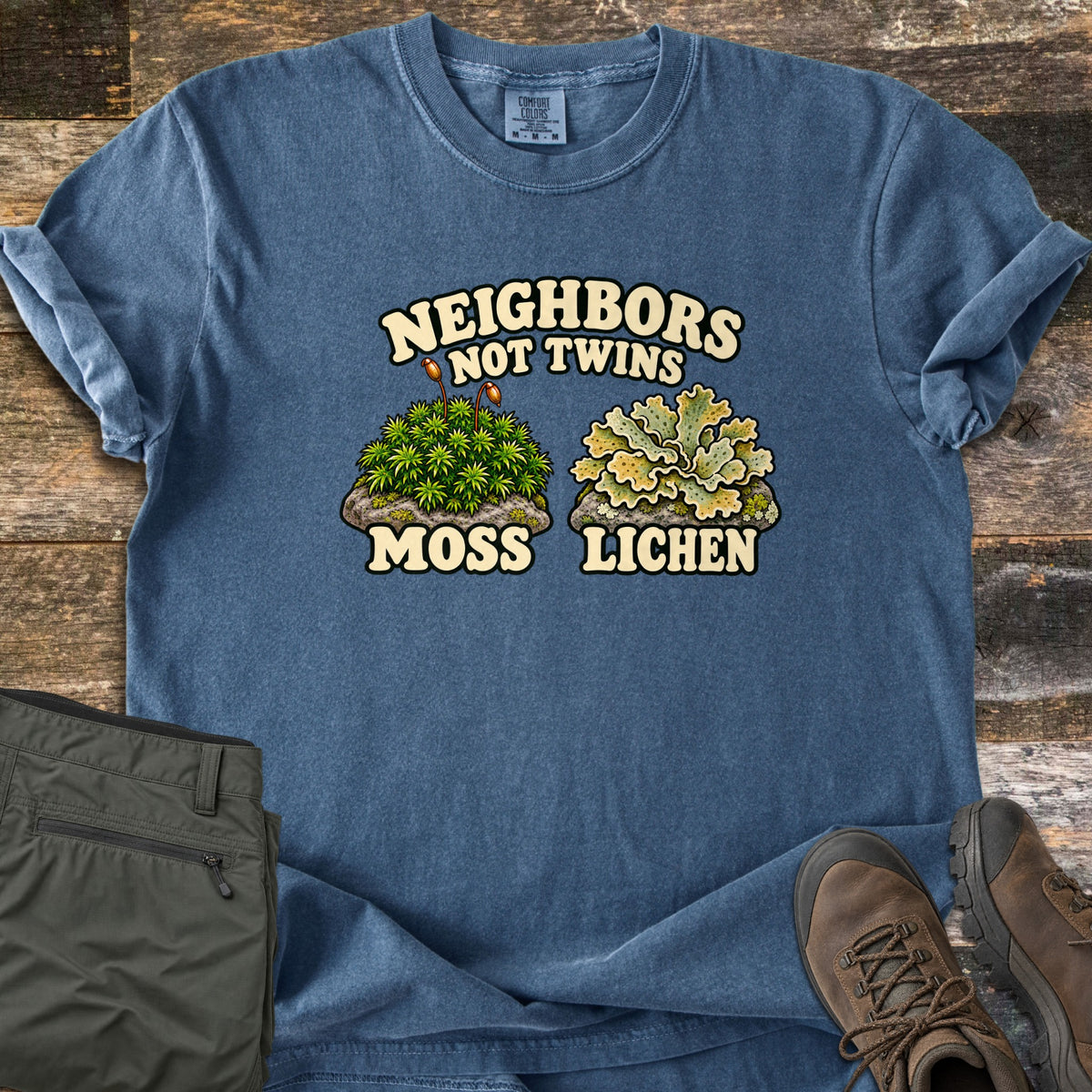 Neighbors Not Twins Comfort Colors T-shirt in Denim, laid flat. Words Neighbors Not Twins over a green moss clump with sporophytes labeled Moss beside a pale leafy lichen labeled Lichen. On weathered wood with olive hiking pants and brown hiking boots.