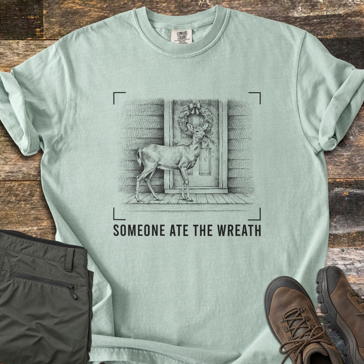 Someone Ate The Wreath Comfort Colors T-shirt in Bay, laid flat. A sketched deer nibbles a wreath on a cabin's front door, framed by corner marks above the words Someone Ate The Wreath. Shown on rustic wood planks with folded olive pants and brown hiking boots.