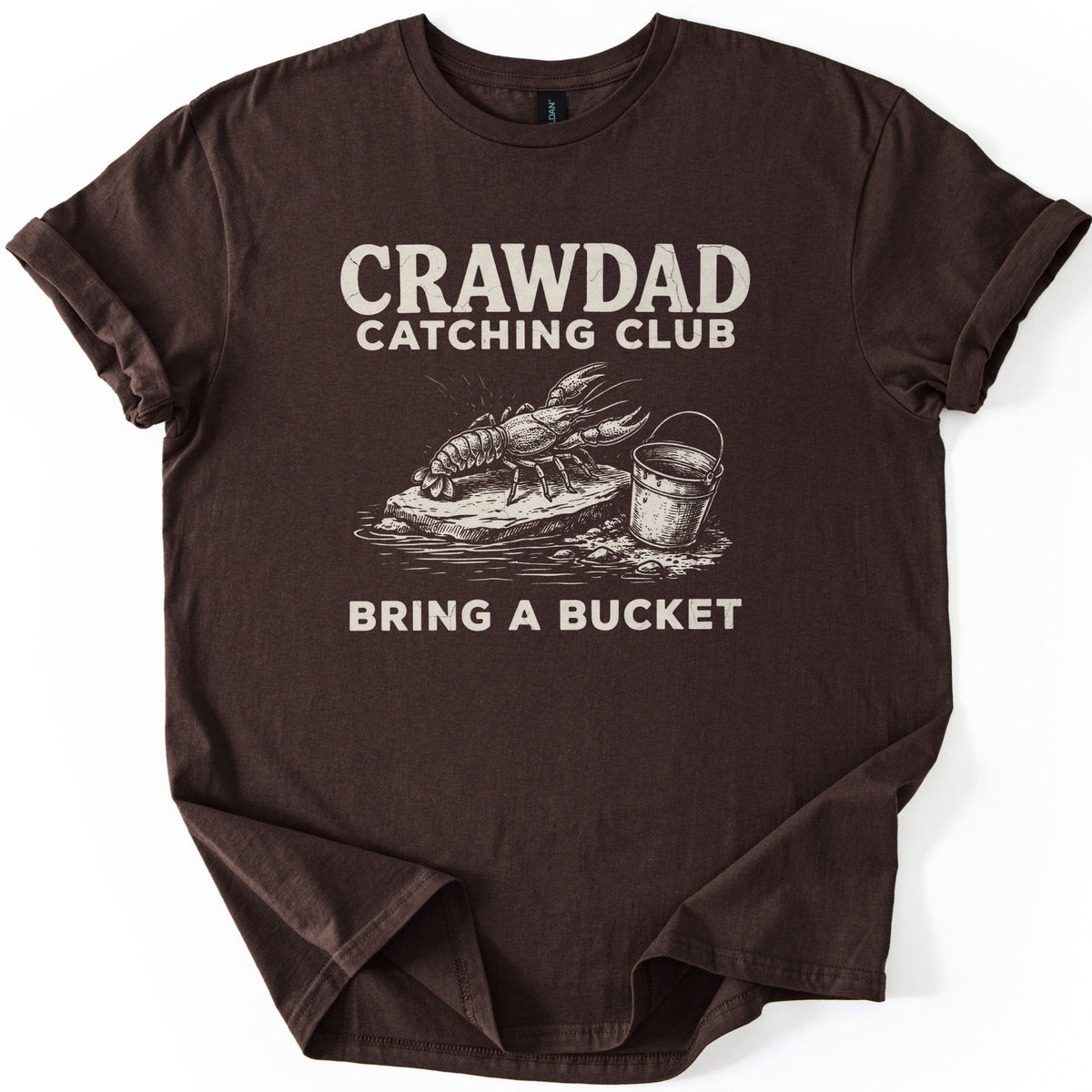 Crawdad Catching Club Comfort Colors T-shirt in Dark Chocolate, laid flat. Cream print reads Crawdad Catching Club above a sketched crawdad on a flat rock at the water's edge beside a metal bucket, with Bring a Bucket below. Cuffed sleeves on a white background.