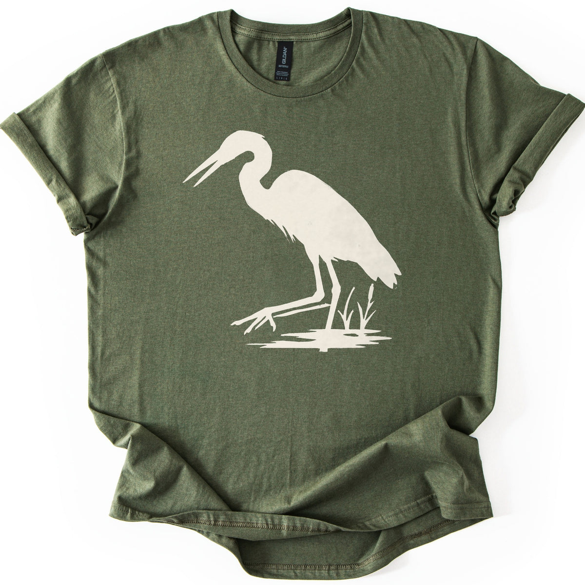 Great Blue Heron Comfort Colors T-shirt in Military Green, laid flat. A cream silhouette of a heron wading in shallow water beside cattails, beak open and one foot reaching forward. The shirt lies on a white surface with cuffed sleeves.