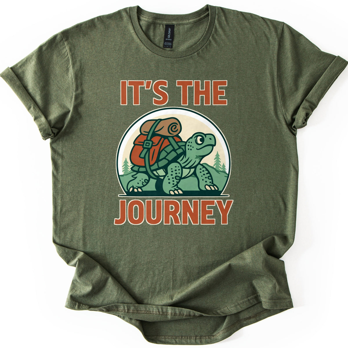 Slow Turtle Hiker Journey Comfort Colors T-shirt in Military Green, laid flat. A green cartoon turtle carrying a red backpack with a rolled sleeping bag walks past pine trees, framed by the words It's the Journey in rust-orange letters. Sleeves are cuffed on a white background.
