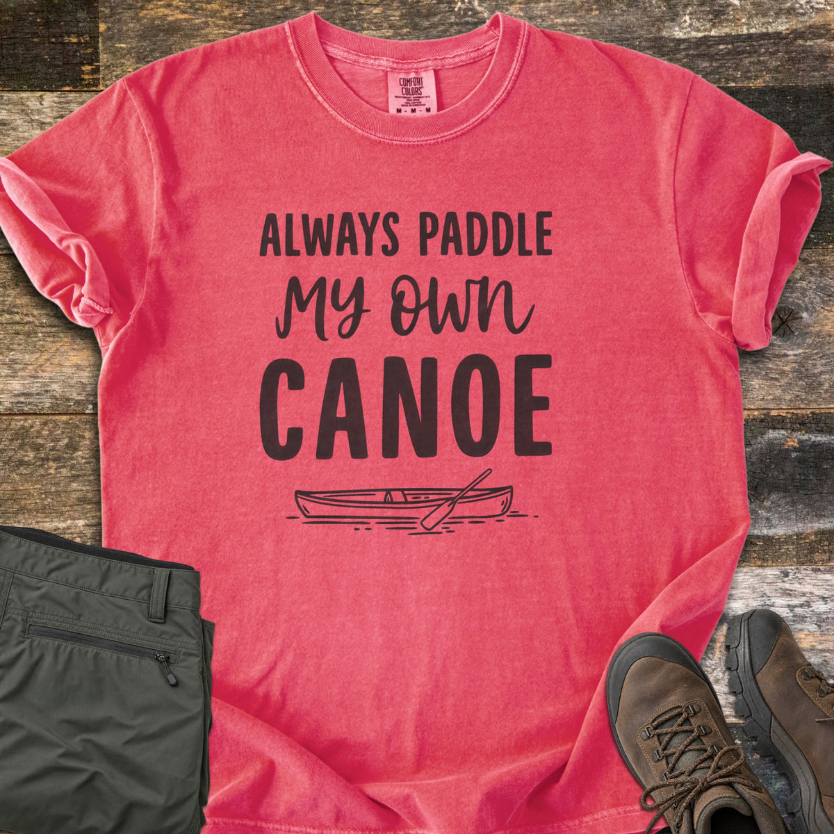 Paddle My Own Canoe Comfort Colors T-shirt in Watermelon, laid flat. Dark text reads Always Paddle My Own Canoe above a line drawing of a canoe with a paddle on the water. It lies on weathered wood with rolled sleeves, folded olive hiking pants, and brown hiking boots.