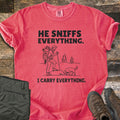 He Sniffs Everything. I Carry Everything Comfort Colors T-shirt in Watermelon, laid flat. Black line art of a hiker with a large backpack and trekking pole beside a dog sniffing a rock, between the words He Sniffs Everything. and I Carry Everything. Olive pants and brown hiking boots sit on weathered wood.