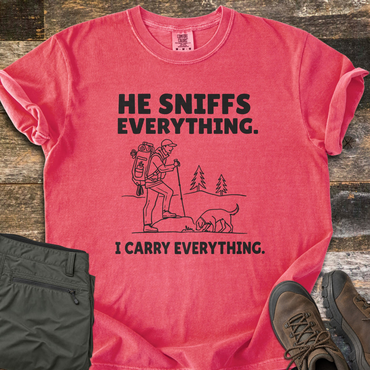 He Sniffs Everything. I Carry Everything Comfort Colors T-shirt in Watermelon, laid flat. Black line art of a hiker with a large backpack and trekking pole beside a dog sniffing a rock, between the words He Sniffs Everything. and I Carry Everything. Olive pants and brown hiking boots sit on weathered wood.