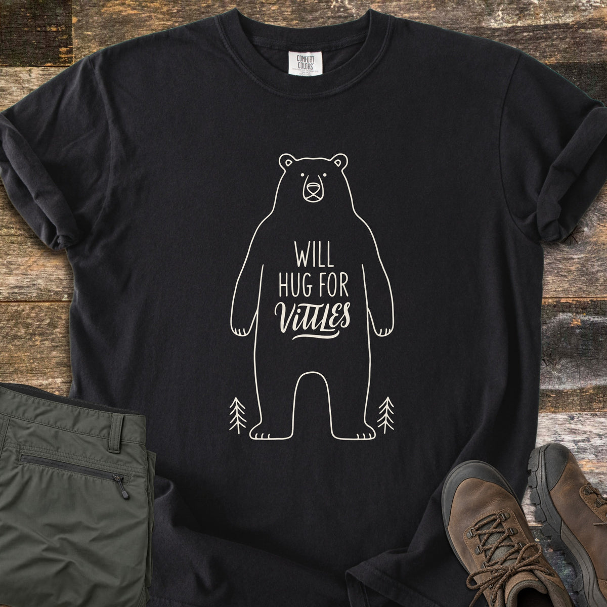 Will Hug For Vittles Comfort Colors T-shirt in Black, laid flat. A cream outline of a standing bear has the words Will Hug For Vittles on its belly, with two small pine trees by its feet. Folded olive hiking pants and brown hiking boots sit beside it on weathered wood.