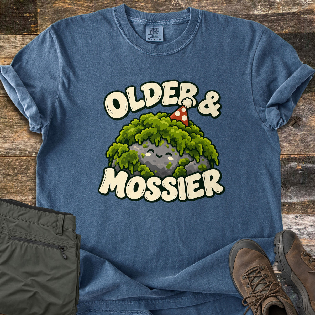 Older & Mossier Comfort Colors T-shirt in Denim, laid flat. A smiling cartoon rock covered in green moss wears a red polka-dot party hat between the words Older & Mossier. The shirt lies on weathered wood with olive hiking pants and brown hiking boots.