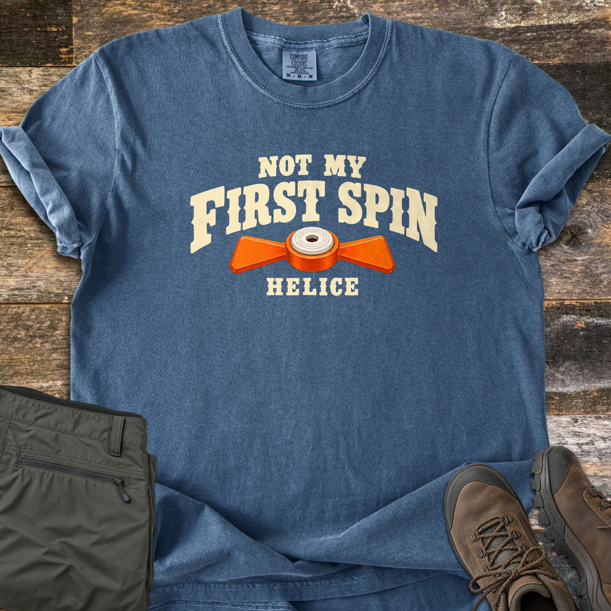 Not My First Spin Comfort Colors T-shirt in Denim, laid flat. Cream arched letters read Not My First Spin above an orange two-winged helice target with a white center, and the word Helice below. Olive pants and brown hiking boots lie beside it on weathered wood planks.