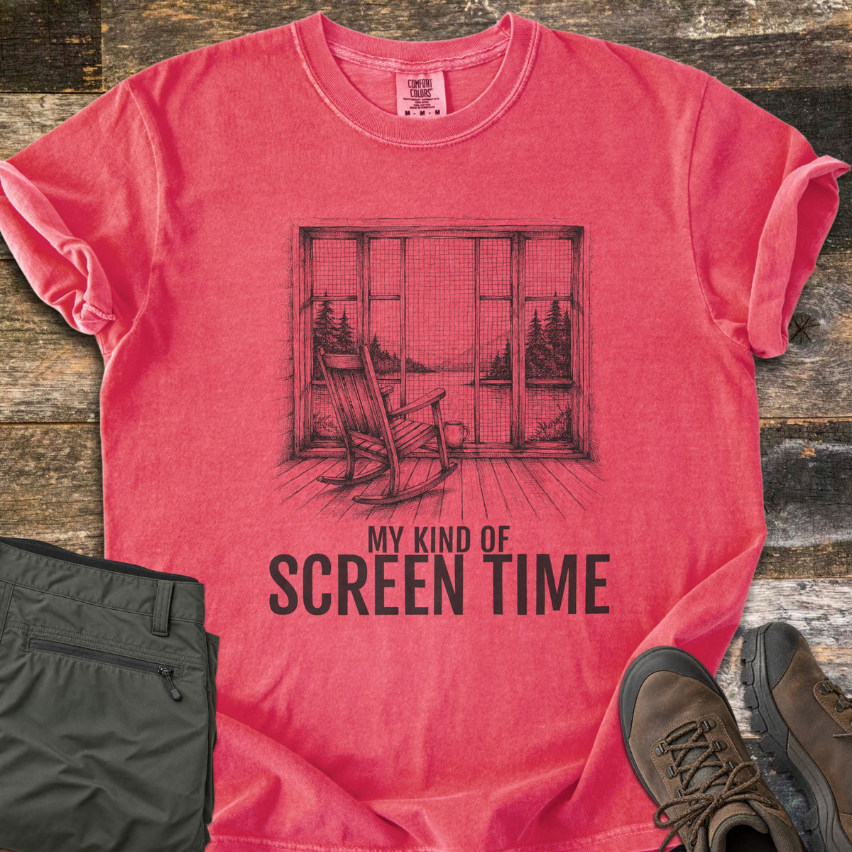 My Kind Of Screen Time Comfort Colors T-shirt in Watermelon, laid flat. A black sketch of a wooden rocking chair and mug facing a screened porch window that overlooks a lake and pine trees, above the words My Kind Of Screen Time. Folded olive hiking pants and brown hiking boots lie on weathered wood.