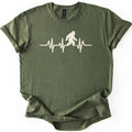 Yeti Heartbeat Comfort Colors T-shirt in Military Green, laid flat. A cream-colored heartbeat line runs across the chest, with a walking Bigfoot silhouette standing within it; there are no words. The sleeves are cuffed and the shirt lies on a white background.