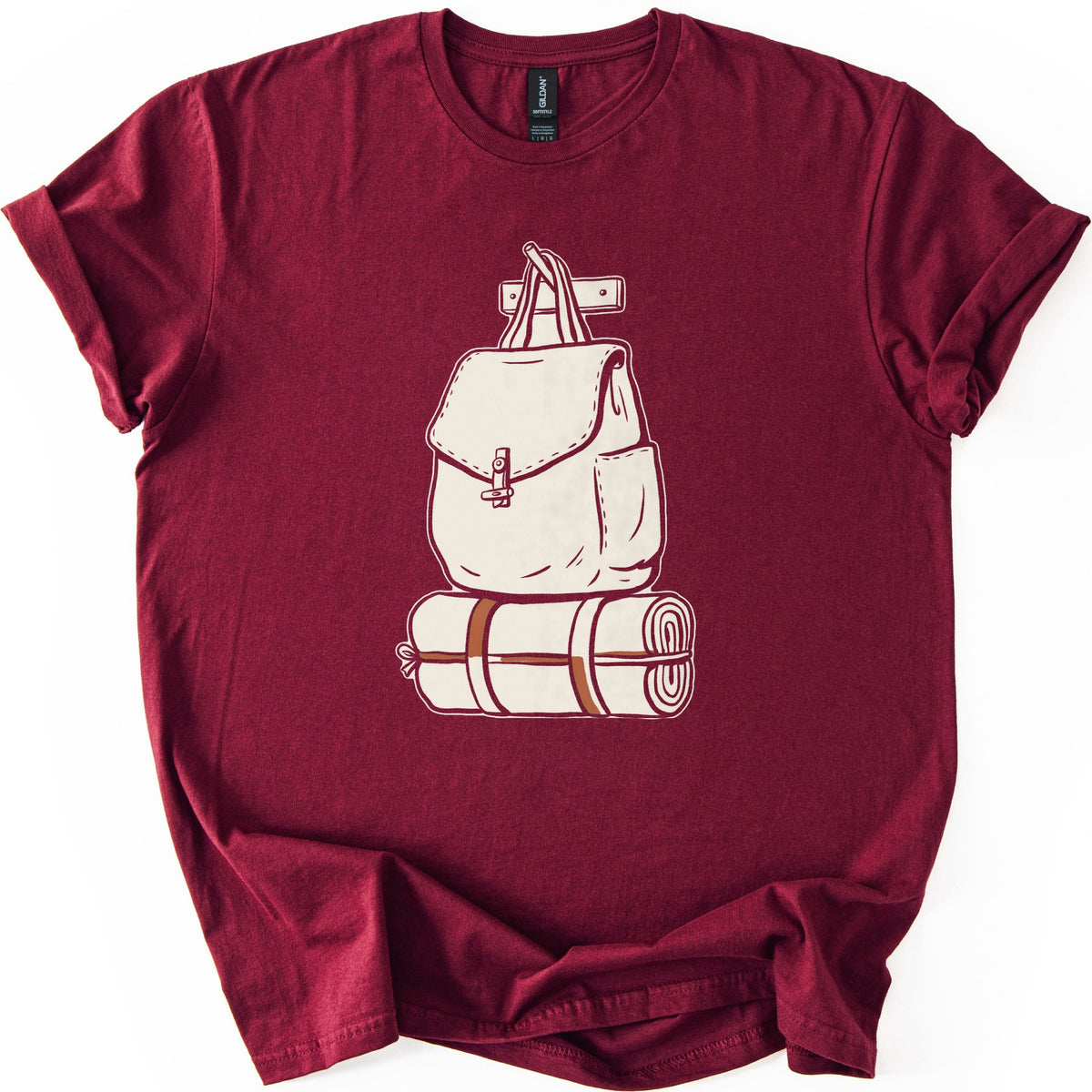 Backpack And Bedroll Comfort Colors T-shirt in Maroon, laid flat. A cream illustration of a canvas backpack with a toggle-clasp flap hangs by its handles from a wall peg above a rolled bedroll tied with rust-brown straps. No words. Cuffed sleeves on a white surface.