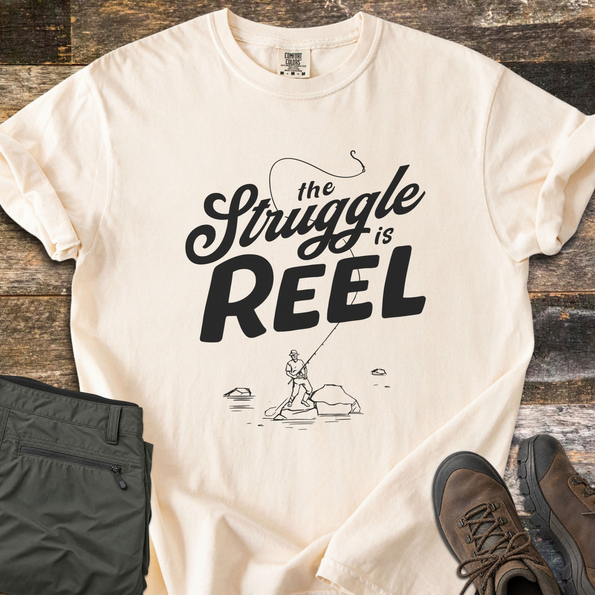 The Struggle Is Reel Comfort Colors T-shirt in Ivory, laid flat. Black text reads the Struggle is REEL under an arcing fishing line with a hook, above a sketch of an angler casting from a rock in water. Folded olive pants and brown hiking boots sit on wood planks.