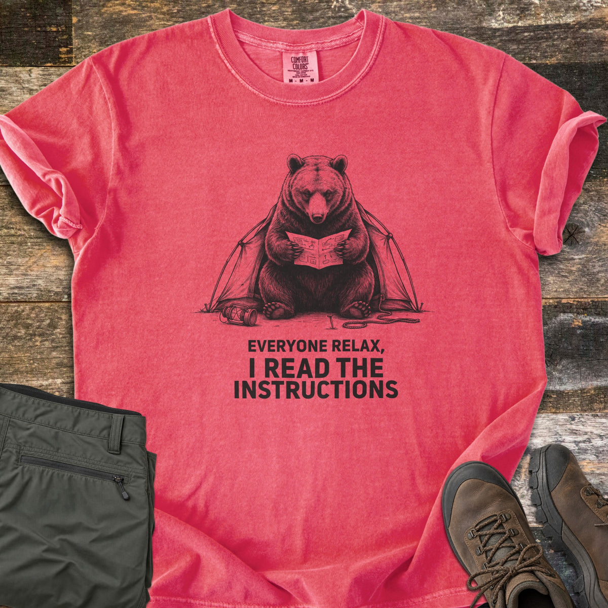 Everyone Relax I Read The Instructions Comfort Colors T-shirt in Watermelon, laid flat. A black sketch of a bear reading instructions in front of a tent, with a tipped lantern and rope, above the words Everyone relax, I read the instructions. Olive cargo pants and brown hiking boots sit on dark wood.