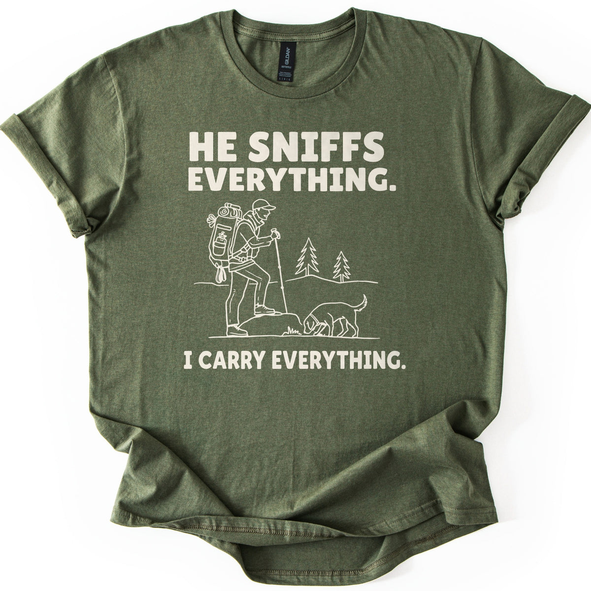 Trail Dog Pack Mule Comfort Colors T-shirt in Military Green, laid flat. Cream line art of a hiker with a loaded backpack and trekking pole stepping onto a rock as a dog sniffs the ground, with pine trees behind, between the words He sniffs everything. and I carry everything. Sleeves rolled, on white.