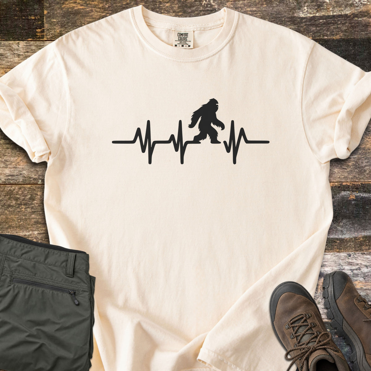 Bigfoot Heartbeat Comfort Colors T-shirt in Ivory, laid flat. A black heartbeat line runs across the chest with a walking Bigfoot silhouette in the middle; there are no words. The shirt lies on weathered wood beside folded olive hiking pants and brown hiking boots.