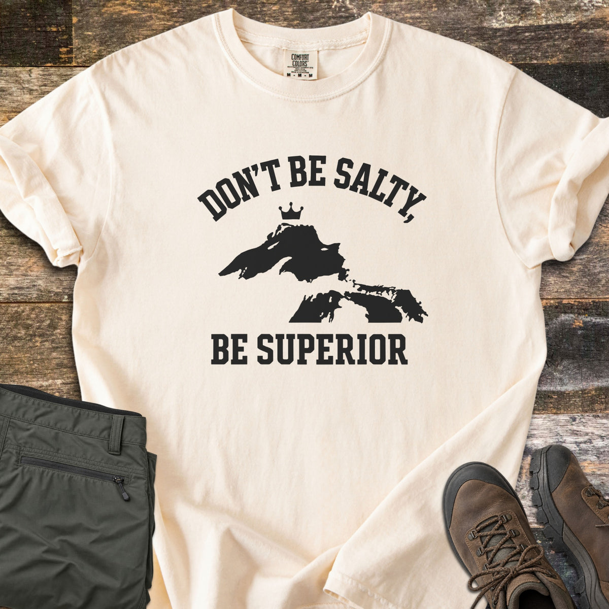 Be Superior Comfort Colors T-shirt in Ivory, laid flat. Black text reads Don't Be Salty, arched over a Lake Superior silhouette topped with a small crown, with Be Superior below. Sleeves rolled, on weathered wood with olive hiking pants and brown hiking boots.