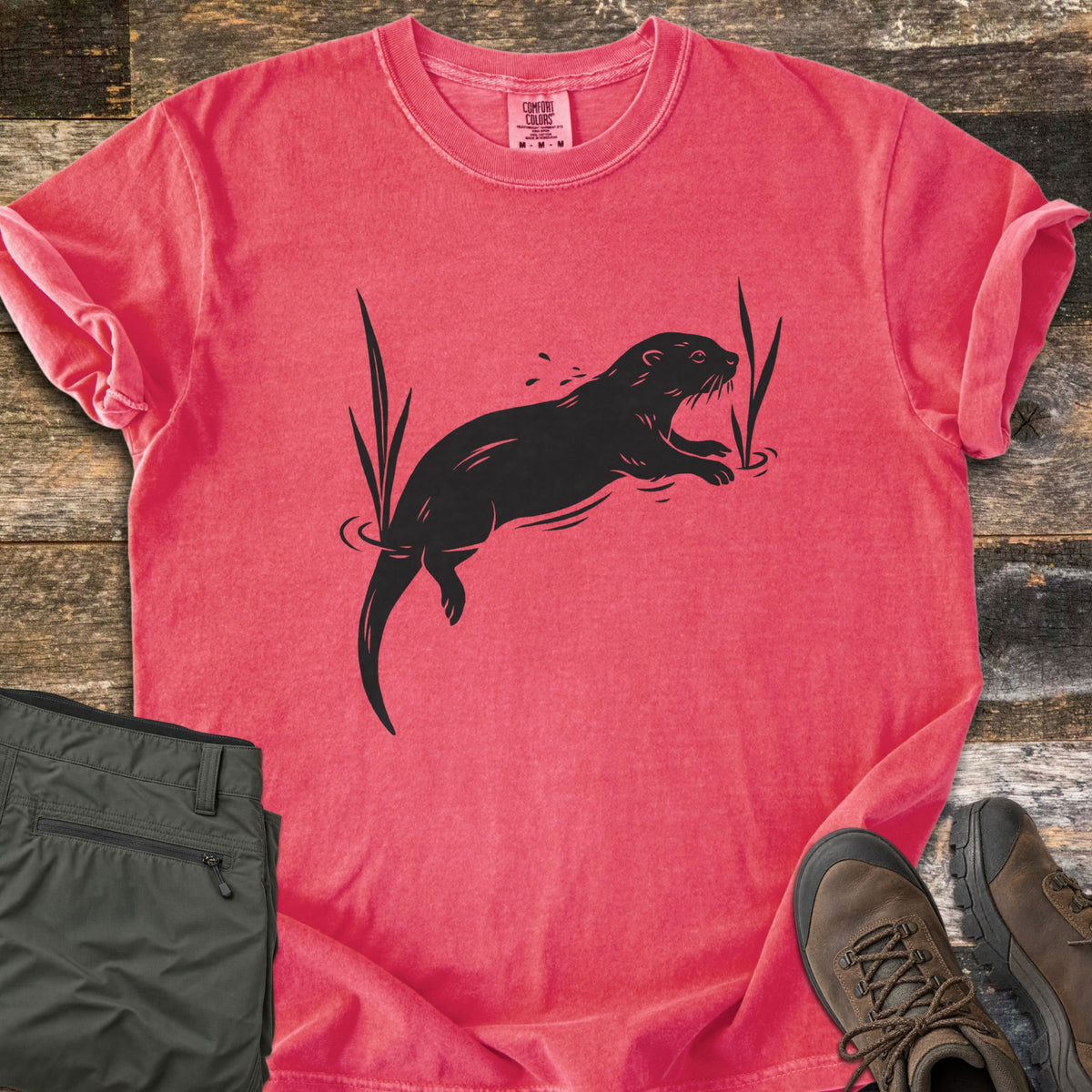 Swimming Otter Comfort Colors T-shirt in Watermelon, laid flat. A black print of an otter swimming between tall reeds, with ripples and splashing droplets. Sleeves are rolled; olive hiking pants and brown hiking boots sit on weathered wood planks.