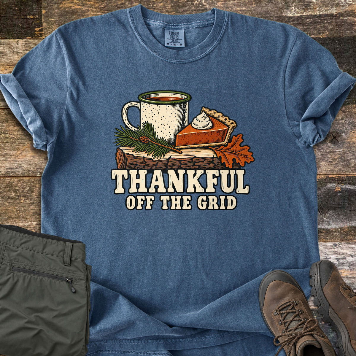 Thankful Off the Grid Comfort Colors T-shirt in Denim, laid flat. A speckled enamel mug of coffee, a slice of pumpkin pie with whipped cream, a pine sprig and an oak leaf sit on a log above the words Thankful Off the Grid. Shown on weathered wood with olive pants and hiking boots.
