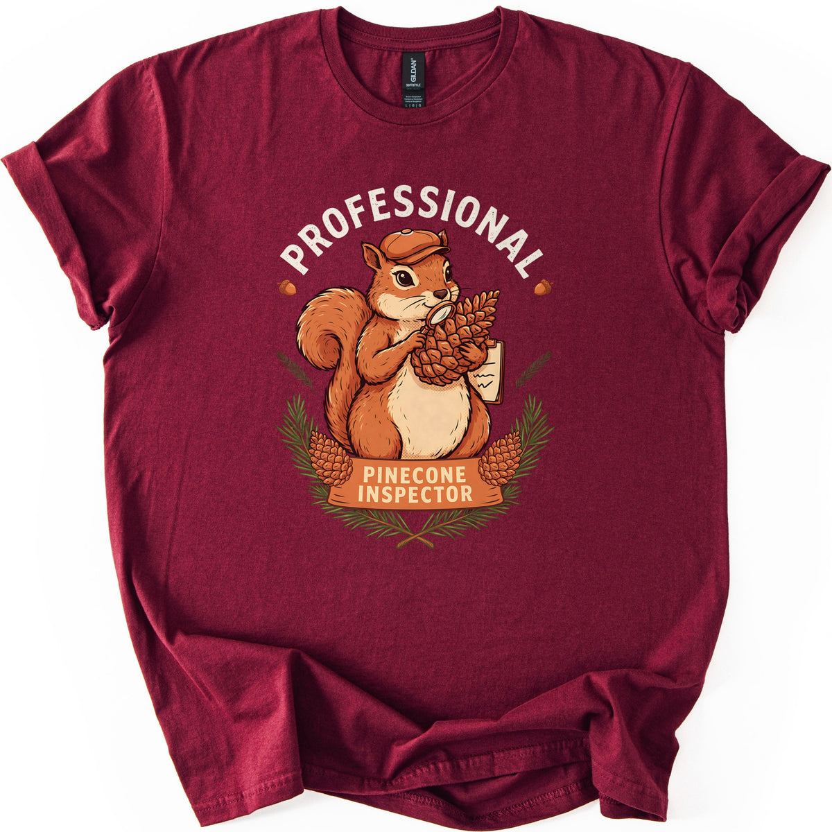 Professional Pinecone Inspector Comfort Colors T-shirt in Maroon, laid flat. A squirrel in a cap inspects a pinecone with a magnifying glass under the arched word Professional in white, above a banner reading Pinecone Inspector framed by pine branches. Sleeves are cuffed on a white surface.