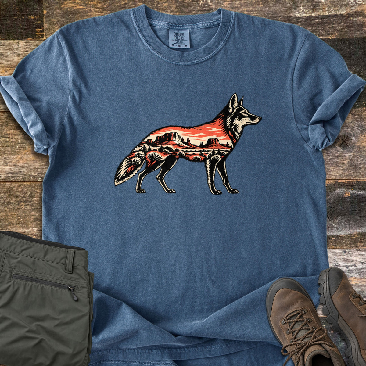 Desert Mesa Coyote Comfort Colors T-shirt in Denim, laid flat. A coyote silhouette in black and cream is filled with an orange desert scene of mesas, buttes and shrubs; no words. Shown on weathered wood with olive pants and brown hiking boots.