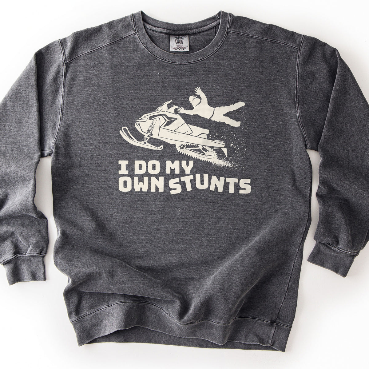 I Do My Own Stunts Comfort Colors crewneck sweatshirt in Pepper, laid flat. A cream print shows a rider in a helmet flying off a snowmobile in midair as snow sprays from the track, above the words I DO MY OWN STUNTS. The sweatshirt lies on a white surface.