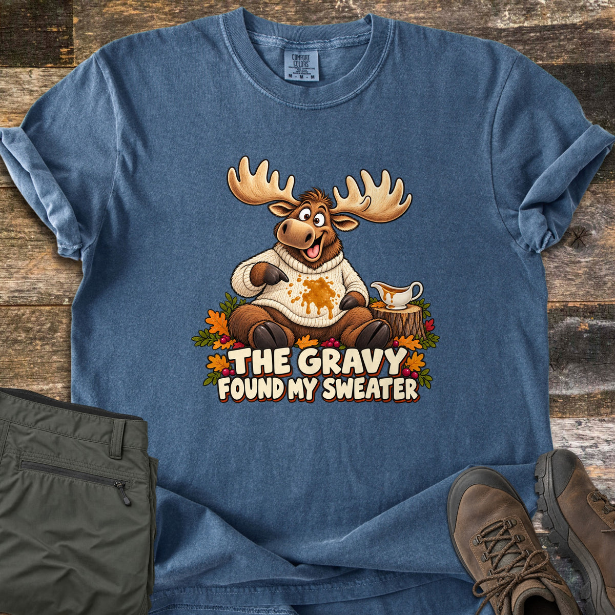 The Gravy Found My Sweater Comfort Colors T-shirt in Denim, laid flat. A cartoon moose in a cream sweater with a gravy stain points at the spill beside a gravy boat on a stump, framed by autumn leaves, above the words The Gravy Found My Sweater. Olive pants and hiking boots sit on wood planks.