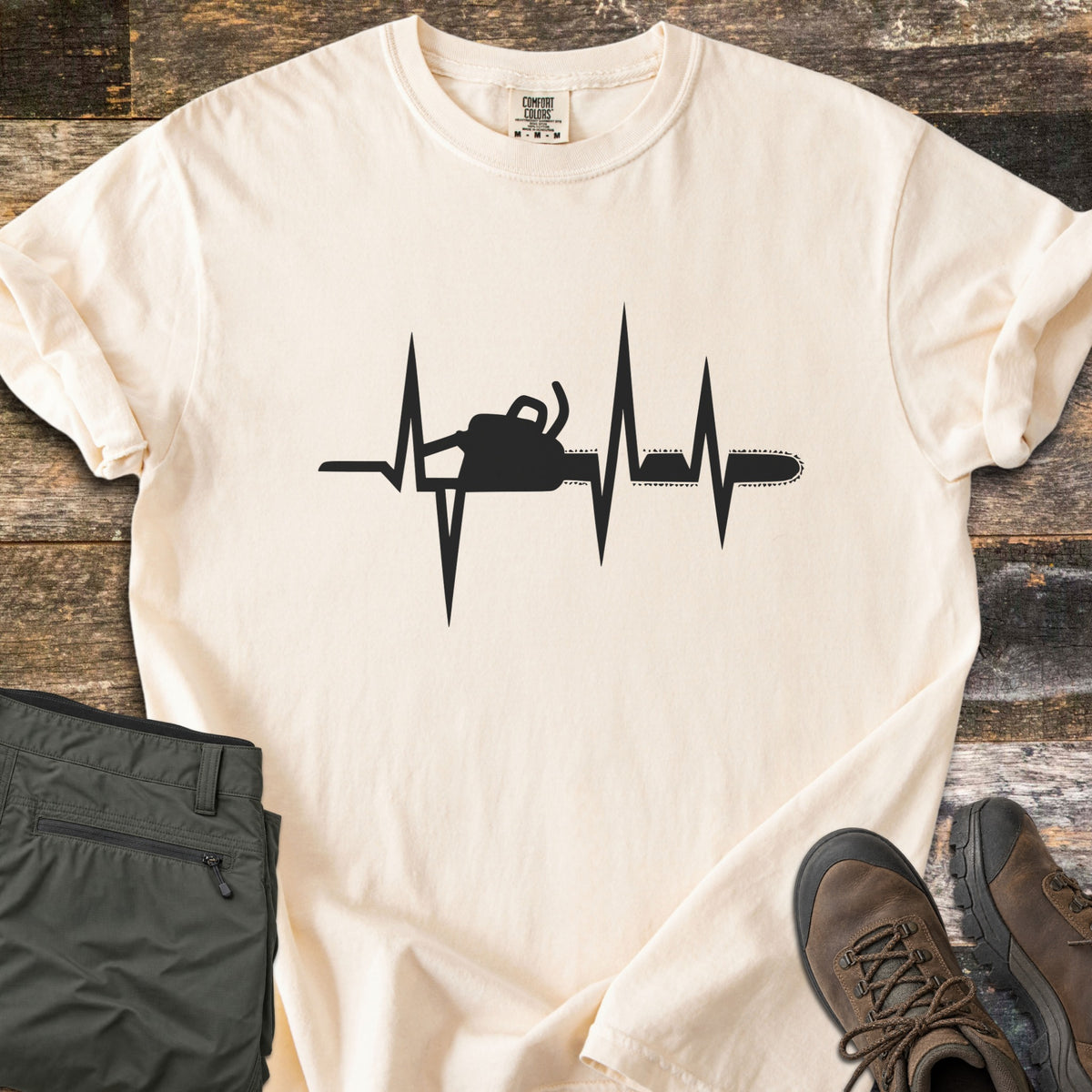 Chainsaw Heartbeat Comfort Colors T-shirt in Ivory, laid flat. A black heartbeat line runs across the chest with a chainsaw silhouette built into it, the bar and chain extending to the right. Folded olive cargo shorts and brown hiking boots sit on a weathered wood floor.